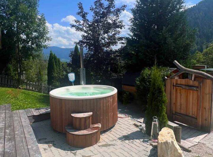 Fasssauna: „Gäste entspannen in der traditionellen Fasssauna im Almdorf Winkl“ Bio-Sauna: „Innenansicht der Bio-Sauna für 8 Personen mit Holzwänden im Almdorf Winkl“ Hot Tub: „Hot Tub im Freien auf der Almwiese mit Bergblick im Almdorf Winkl“