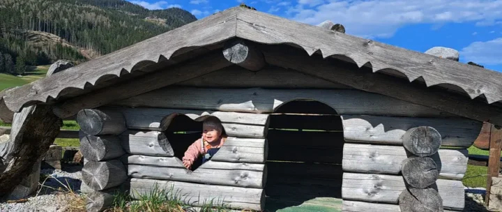 Abenteuerspielplatz: „Kinder auf dem Abenteuerspielplatz mit Rutsche und Klettergerüst im Almdorf Winkl“ Trampolin: „Gäste springen auf dem großen Trampolin inmitten der Alm Wiese“ Lagerfeuerplatz: „Lagerfeuerstelle mit Holzstühlen und Grillrost im Almdorf Winkl“