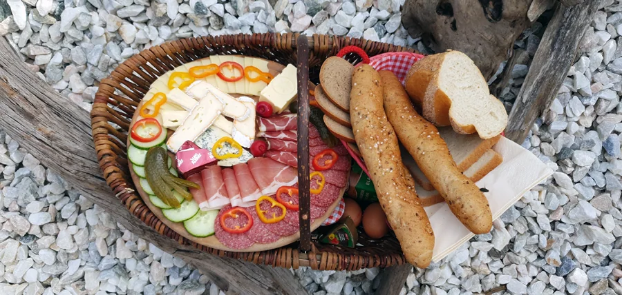 Frühstückskorb Gesamt: „Liebevoll gefüllter Frühstückskorb mit Wurst, Käse und Marmelade auf Holztisch im Almdorf Winkl“ Steierkas: „Nahaufnahme des regionalen Steierkas im Frühstückskorb“ Marmeladenauswahl: „Hausgemachte Erdbeer-, Himbeer- und Aprikosenmarmeladen im Almdorf Winkl“